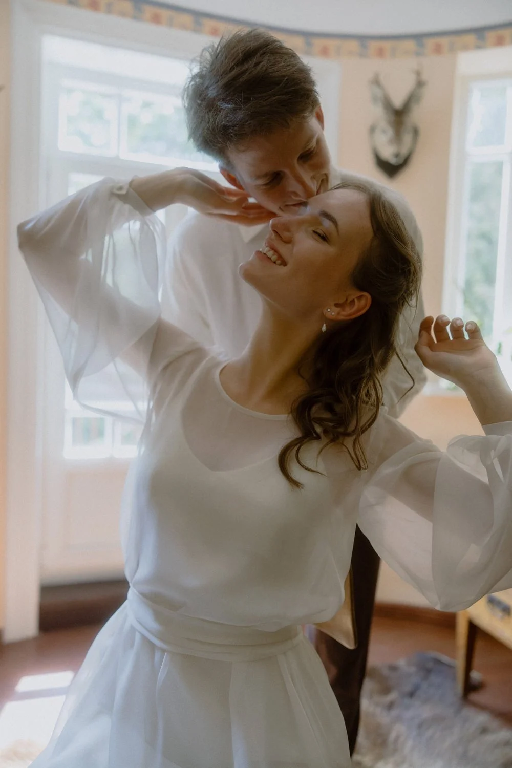 A couple smiling and embracing indoors near windows, with the woman wearing a white dress and the man leaning over her.