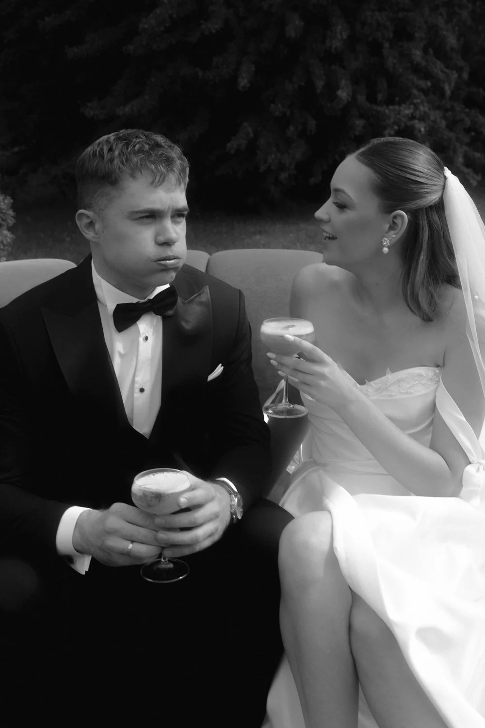 Black and white photo of a bride and groom sitting outdoors. The groom is looking away with a serious expression, wearing a tuxedo, and holding a cocktail glass. The bride is smiling, wearing a wedding dress and veil, and holding a cocktail glass, se
