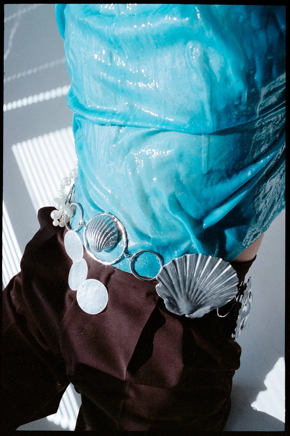 A person wearing a large blue, shiny, textured head covering with shells and pearl jewelry attached, dressed in dark clothing, standing in a space with light and shadow patterns.