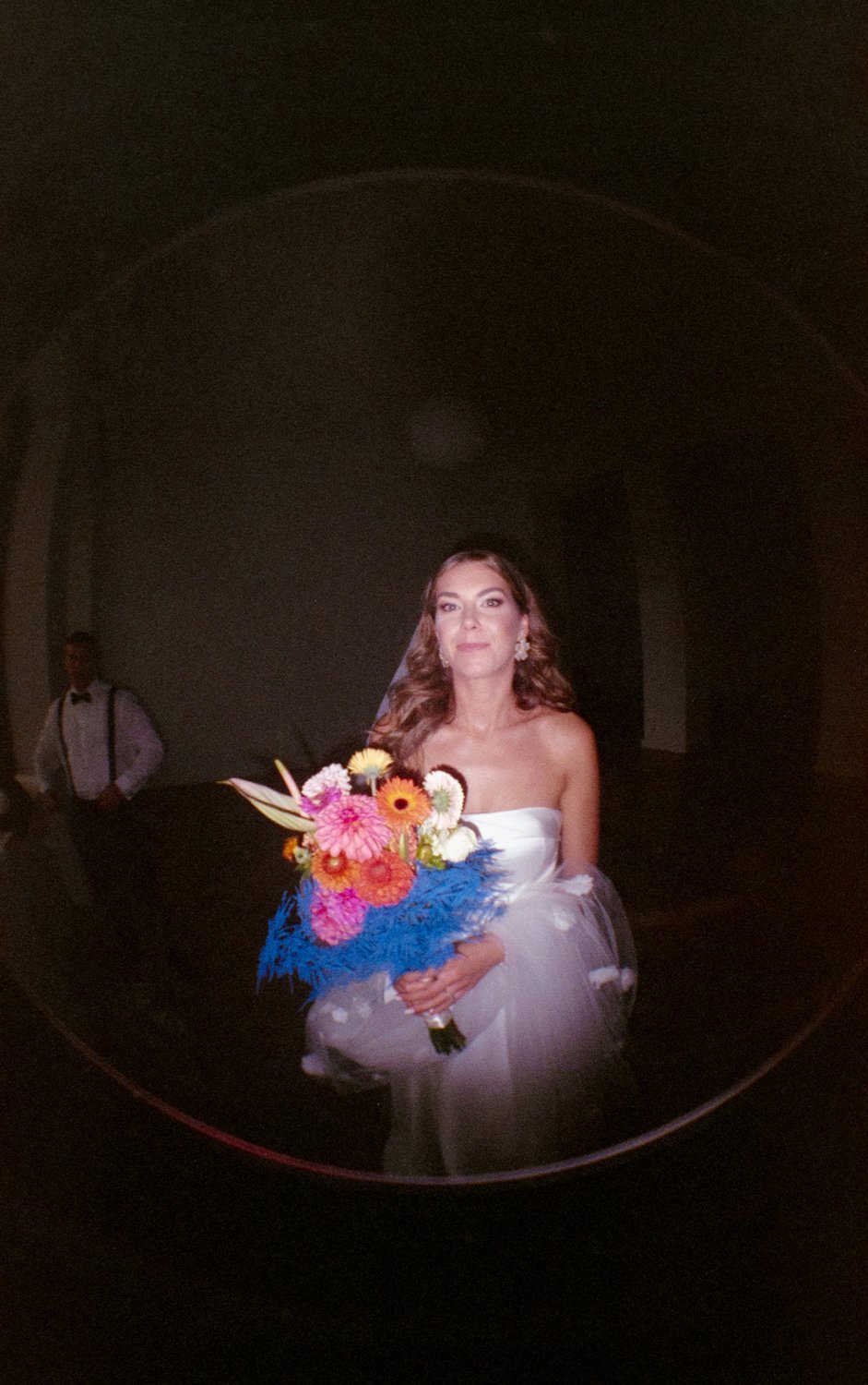 A woman in a white strapless gown holding a colorful bouquet of flowers, standing in a dimly lit room.