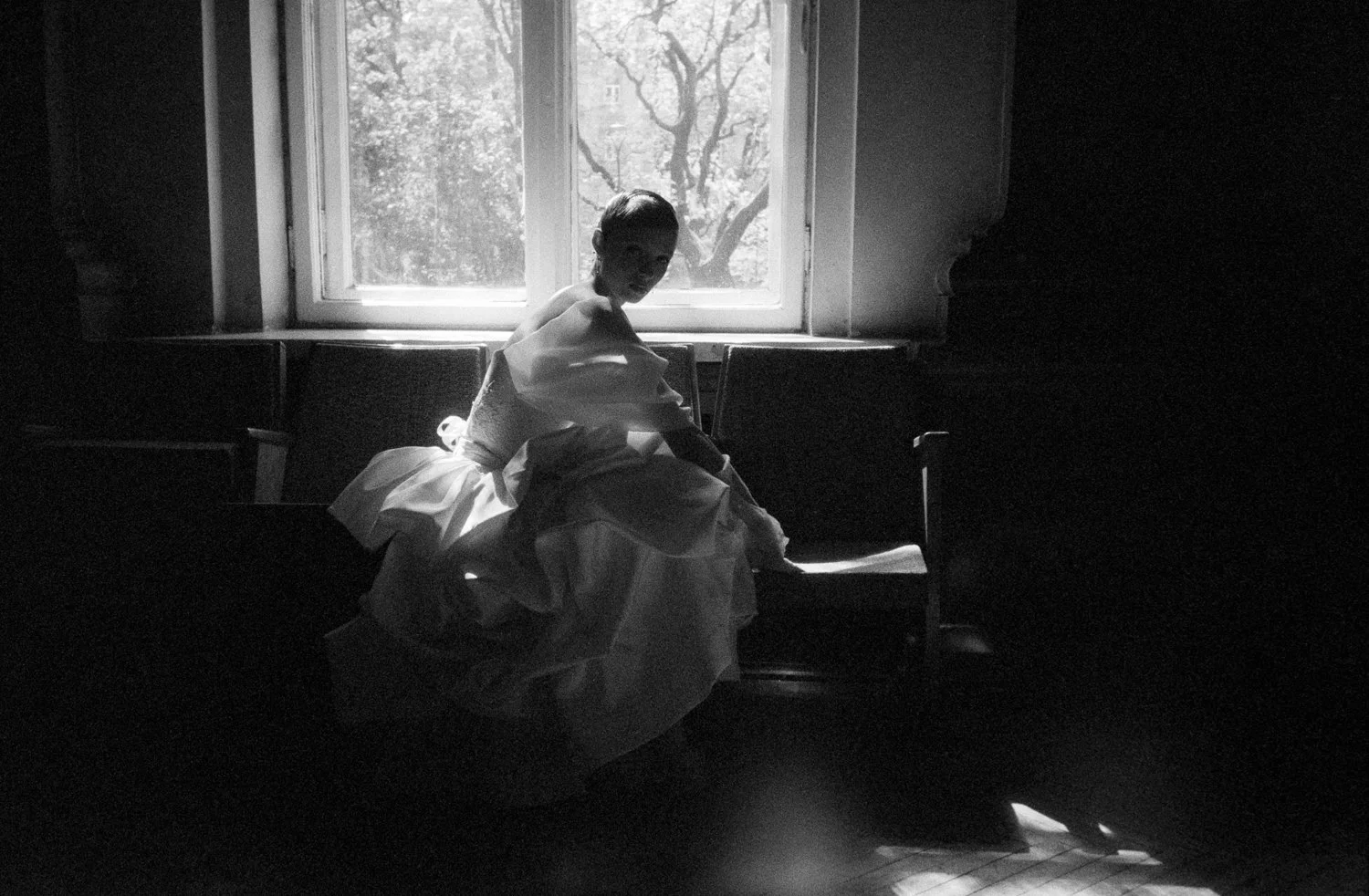 A woman in a white dress sitting on a bench near a window, with light streaming in and trees visible outside.