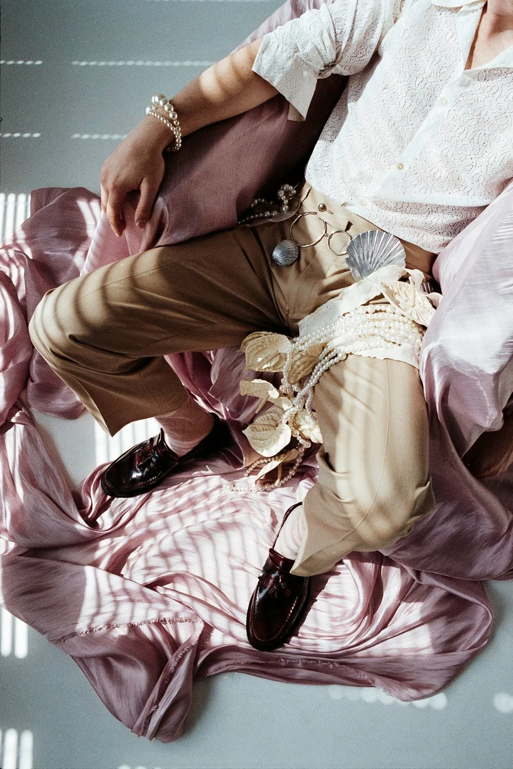 Close-up of a person wearing beige pants, a white lace shirt, and dark shoes, sitting on pink satin fabric, with accessories like pearls, shells, and rings.