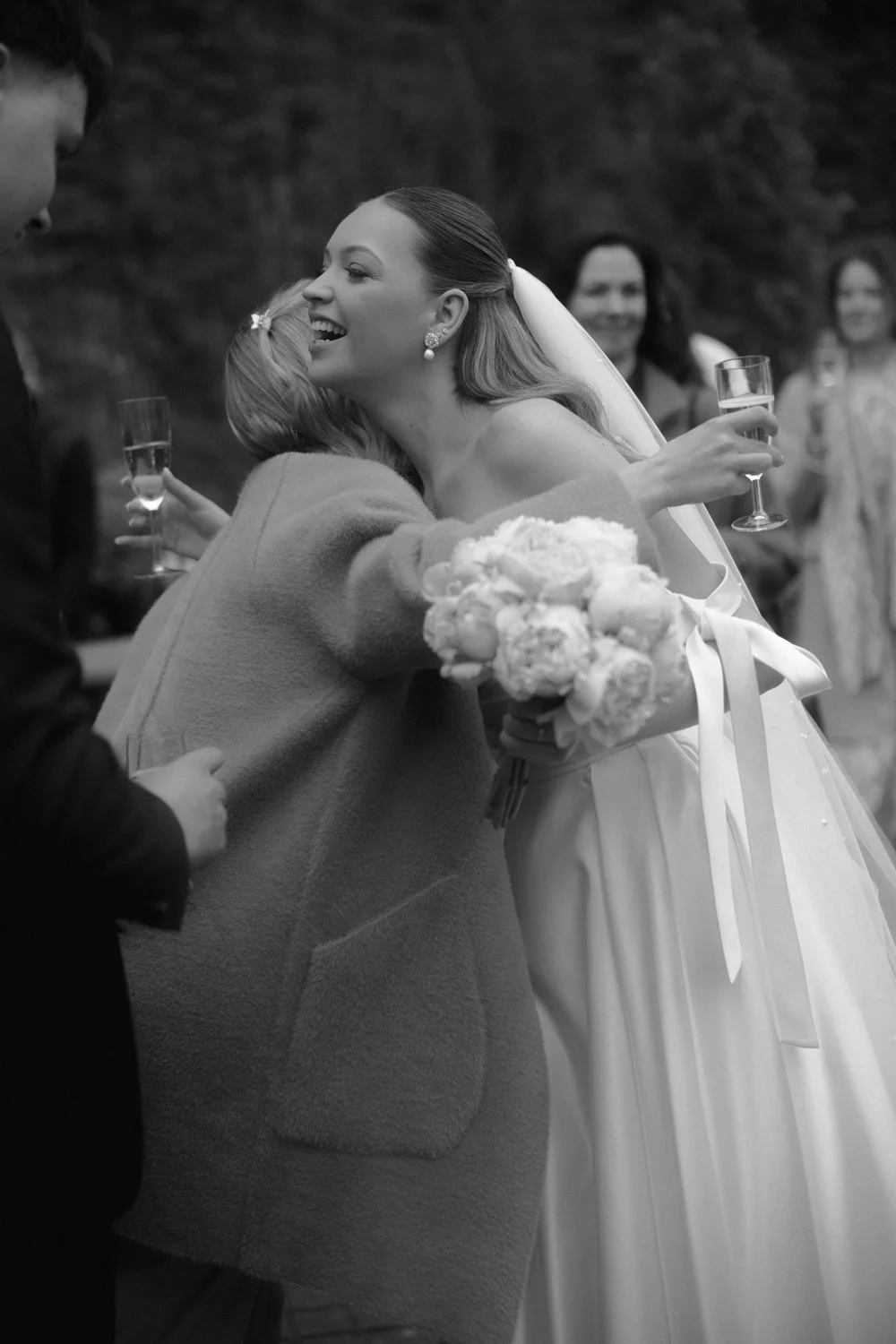 A woman in a wedding dress is hugging a guest during a celebration, holding a bouquet of flowers and a glass of champagne, with other guests in the background.