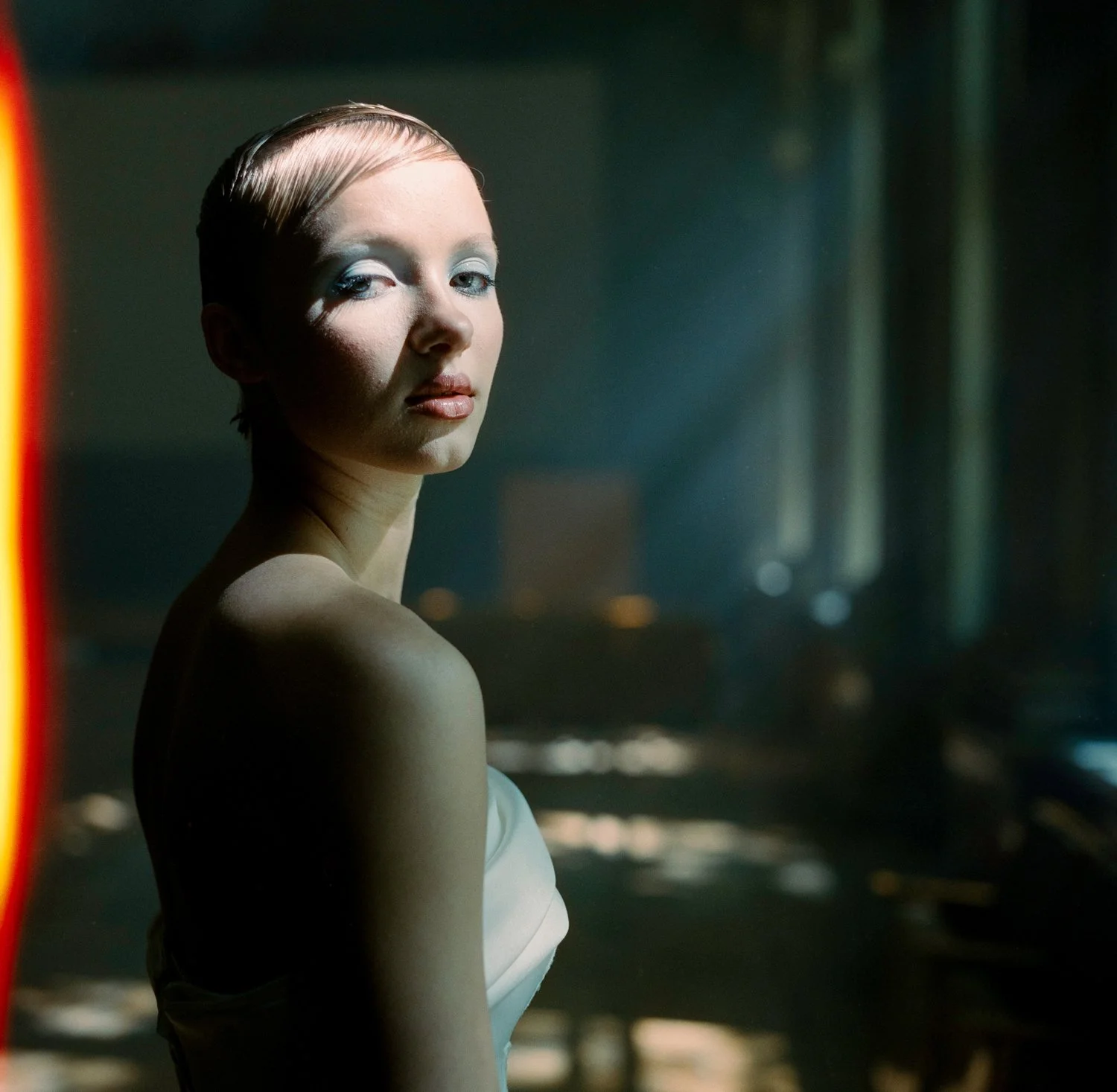 A woman with short, slicked-back hair and light makeup, wearing a strapless white dress, standing in a dimly lit room with a cityscape in the background.