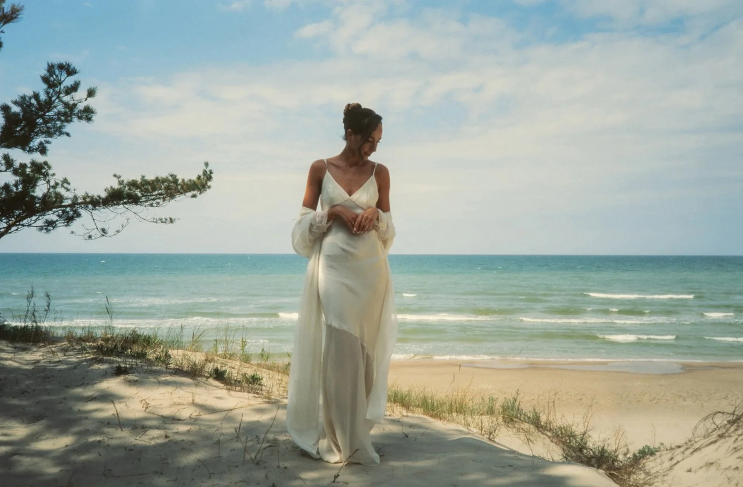 A woman in a white dress standing on a sandy beach near the ocean with a tree on the left side and a partly cloudy sky.
