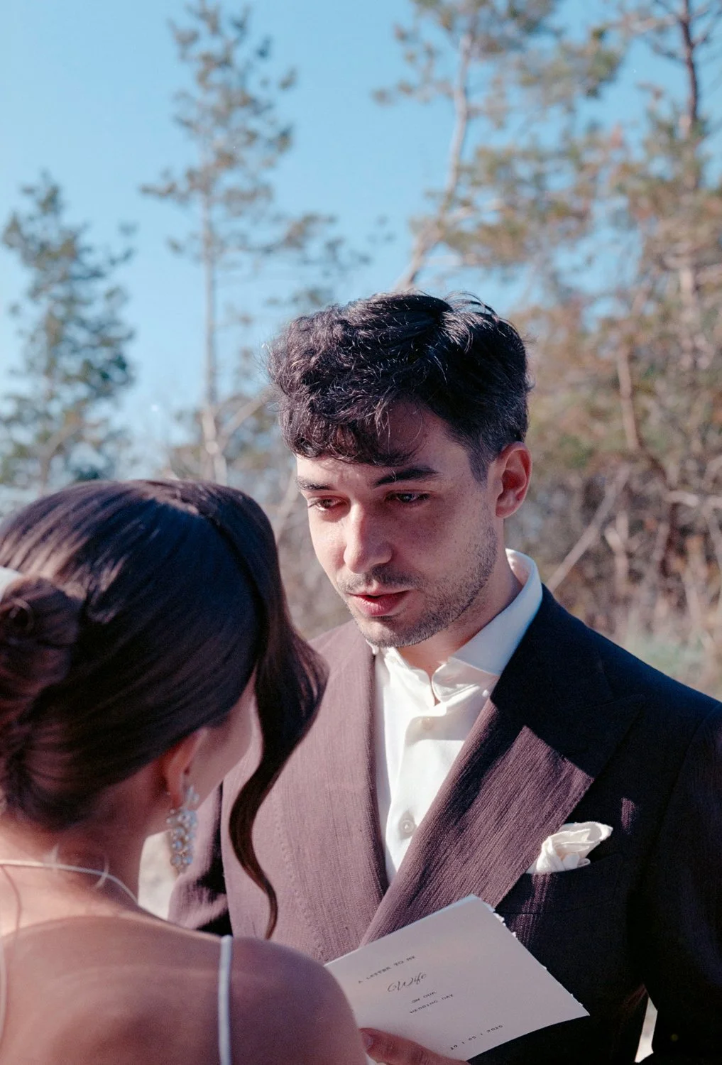 A man and woman at a wedding ceremony, with the woman holding a wedding program while they exchange vows outdoors during daytime.