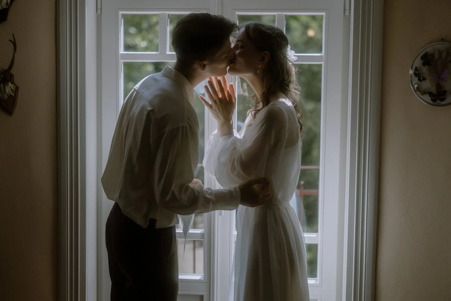 A couple kissing in front of a window, backlit by natural light, with trees visible outside.