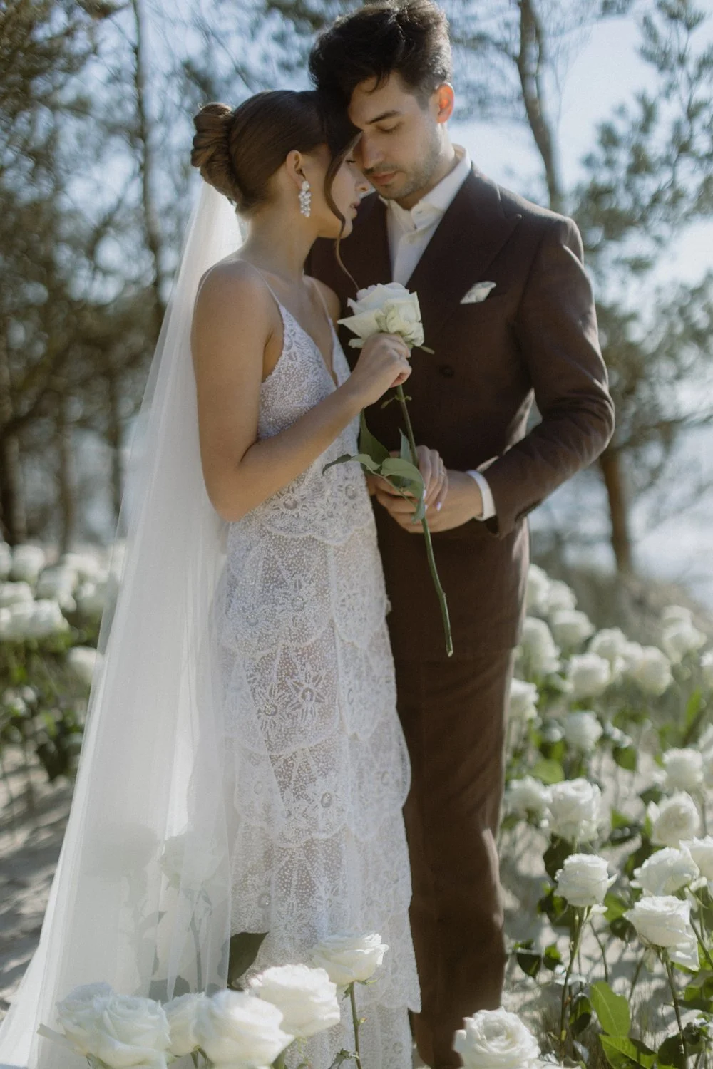 A bride and groom standing close together outdoors in a garden surrounded by white roses, with the groom holding a white rose.