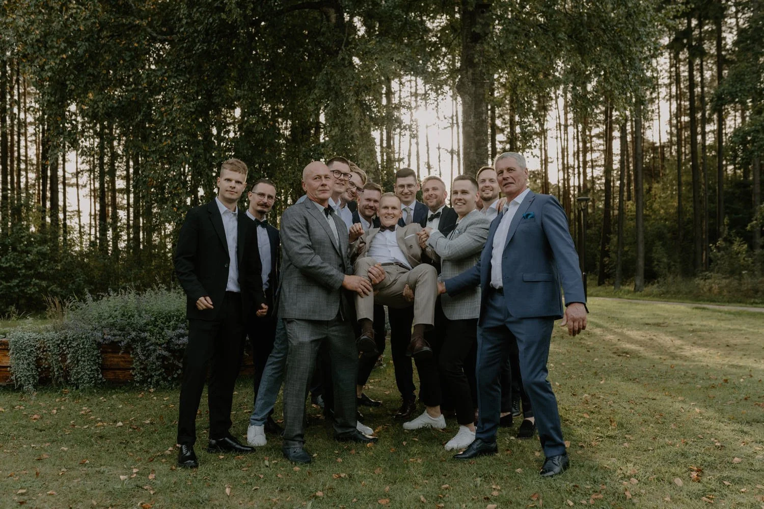 A group of men dressed in formal suits gathers outdoors in a forested area with tall trees and green grass. One man is being held up and carried by others, smiling and enjoying the moment during a celebration, possibly a wedding or similar event.