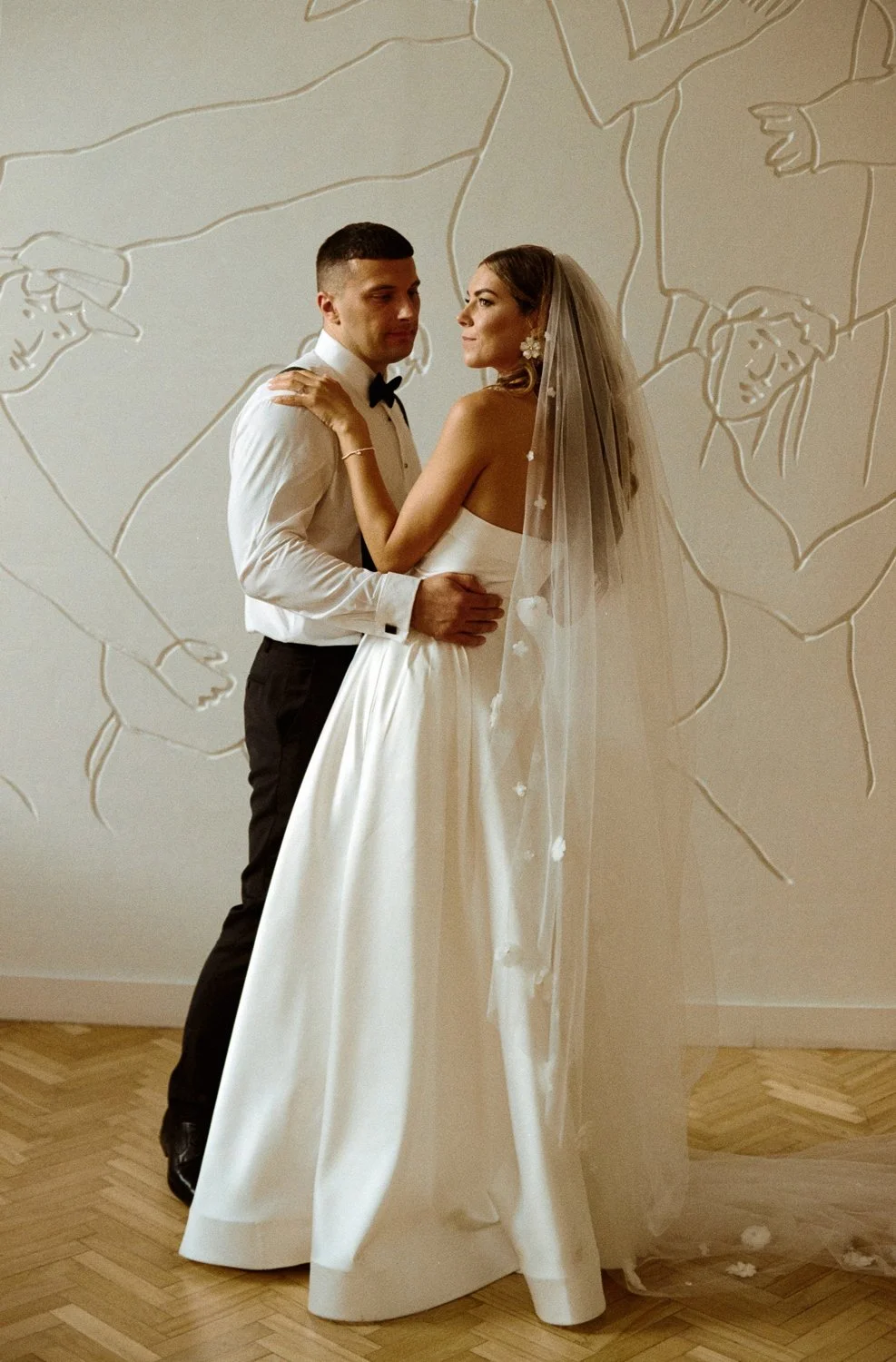 A bride and groom posing together indoors, with a wall featuring artistic line drawings in the background.
