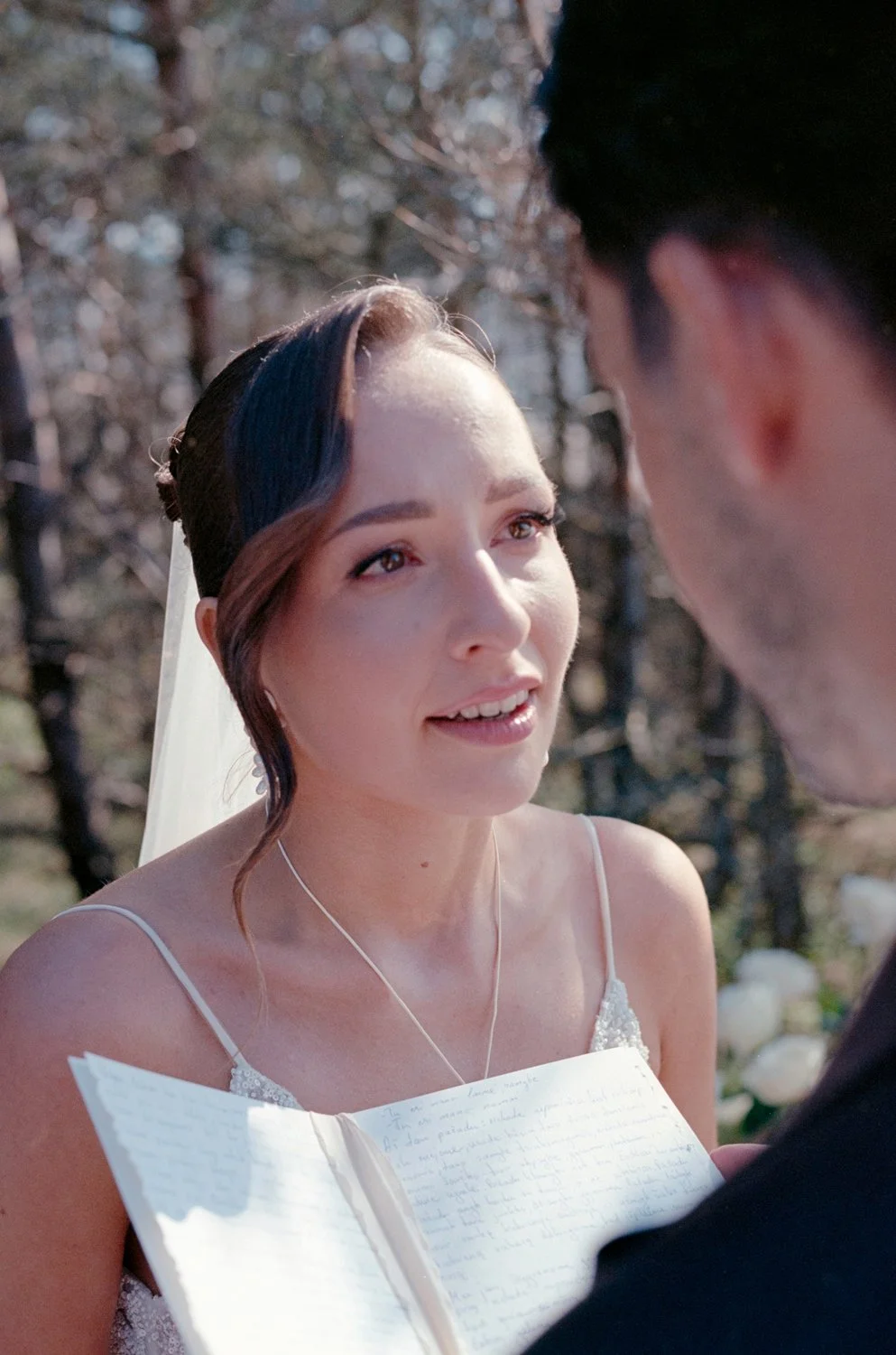A bride with dark hair, wearing a white wedding dress and a necklace, looks at a groom holding an open notebook outdoors during a wedding ceremony.