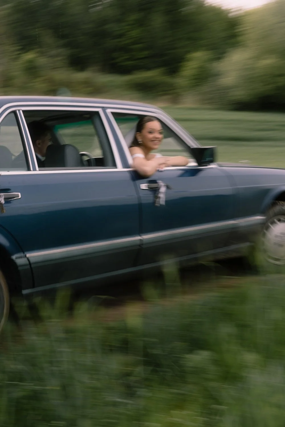 A woman smiling and leaning out of a moving blue car’s window, with blurred greenery in the background.