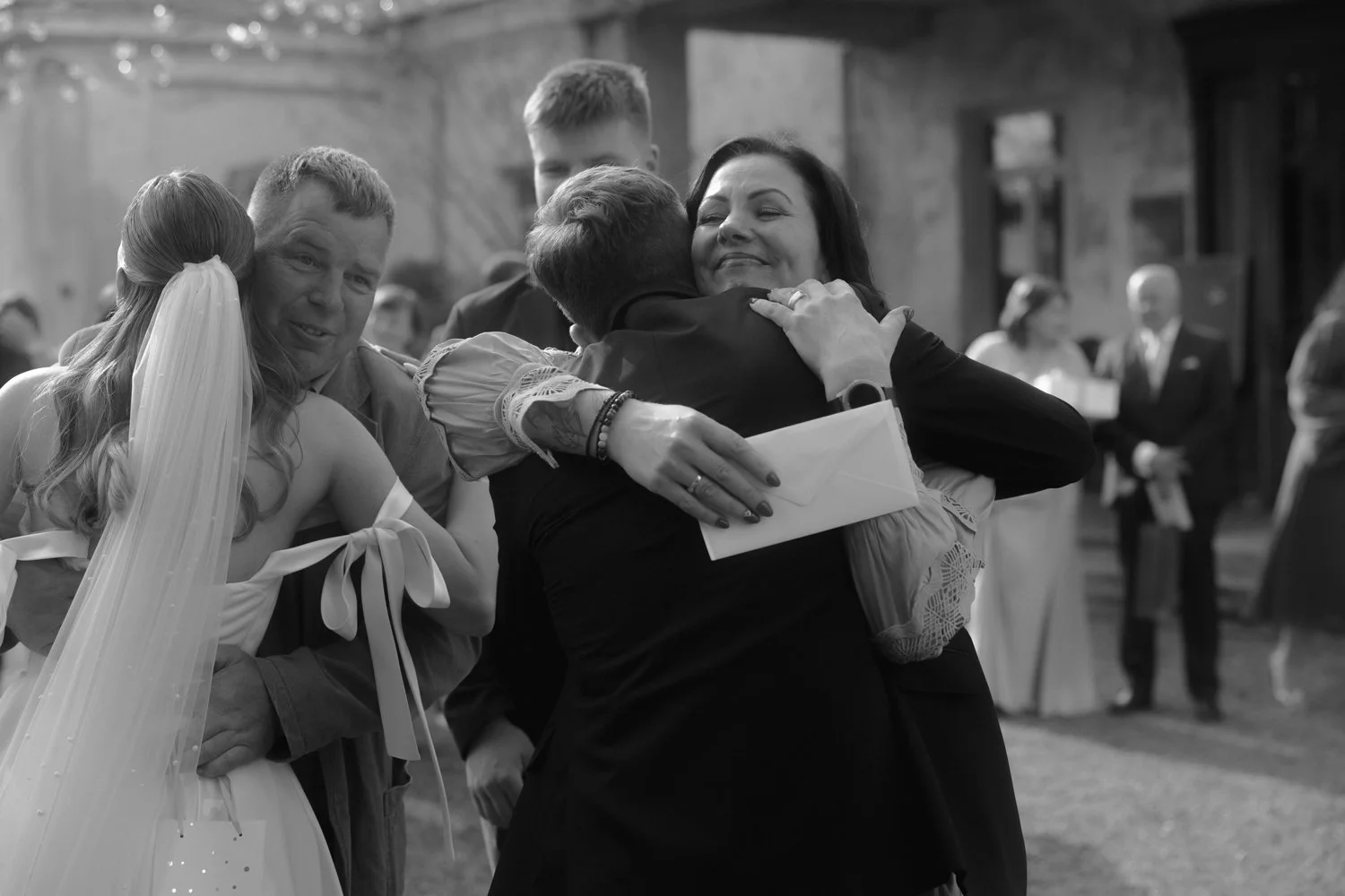 People hugging and smiling at a wedding reception, with others in the background.