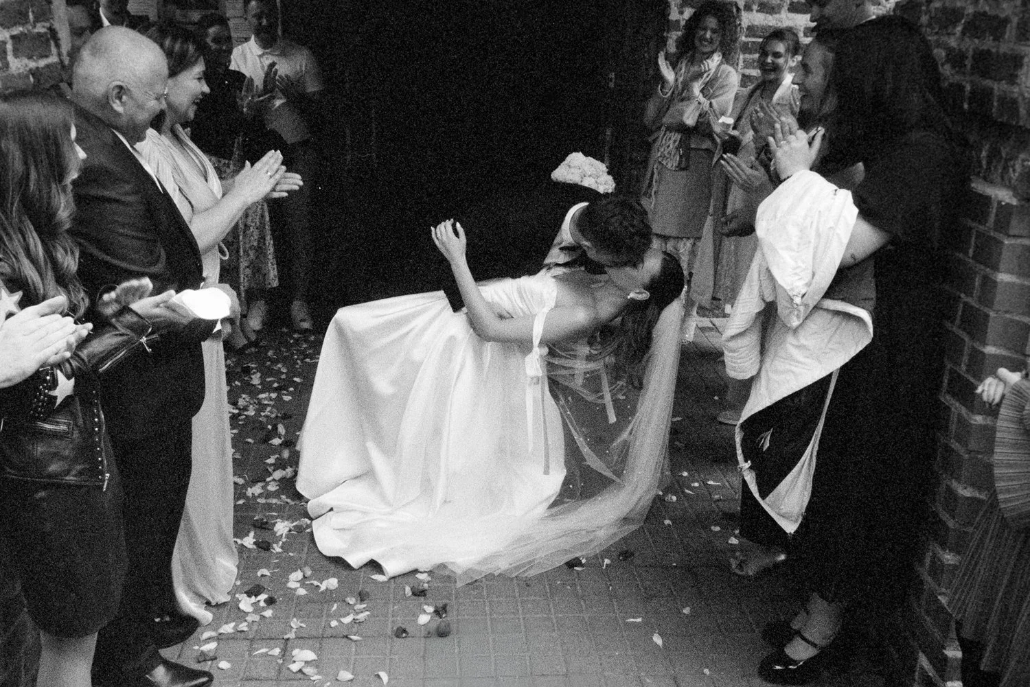 A couple sharing a kiss at their wedding reception, with the bride in a white wedding dress and the groom in formal attire. Guests surround them, clapping and smiling, as flower petals are scattered on the floor.