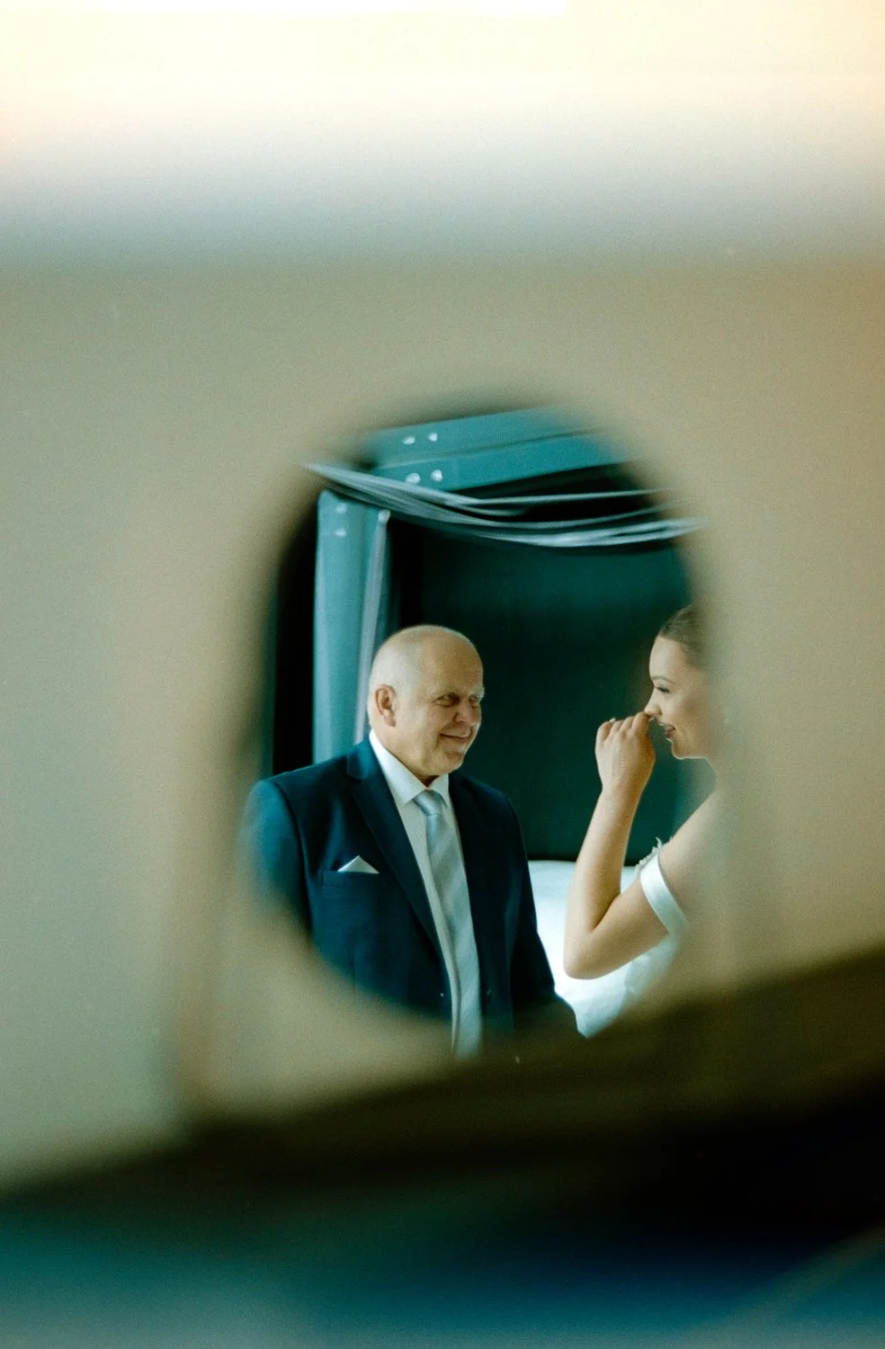 A view through a peephole shows a man in a suit smiling and talking to a woman, who appears to be touching her nose, in a room with a bed and stacked pillows.