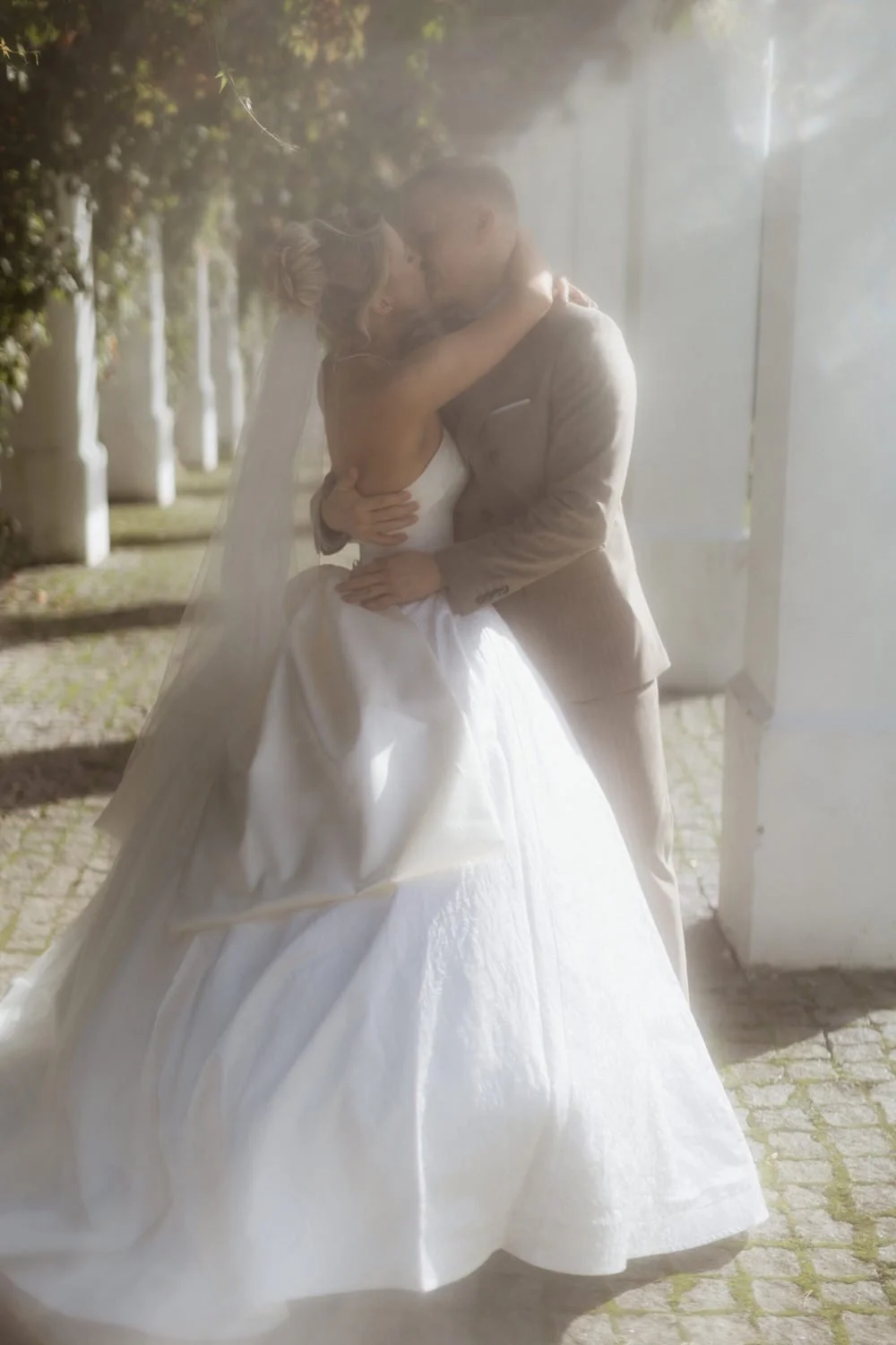 A bride and groom sharing a kiss outdoors, with the bride in a white wedding gown and the groom in a beige suit, surrounded by greenery.