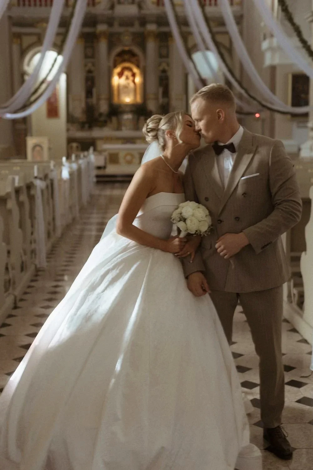 A bride and groom sharing a kiss inside a decorated church, with the bride holding a bouquet of white flowers.