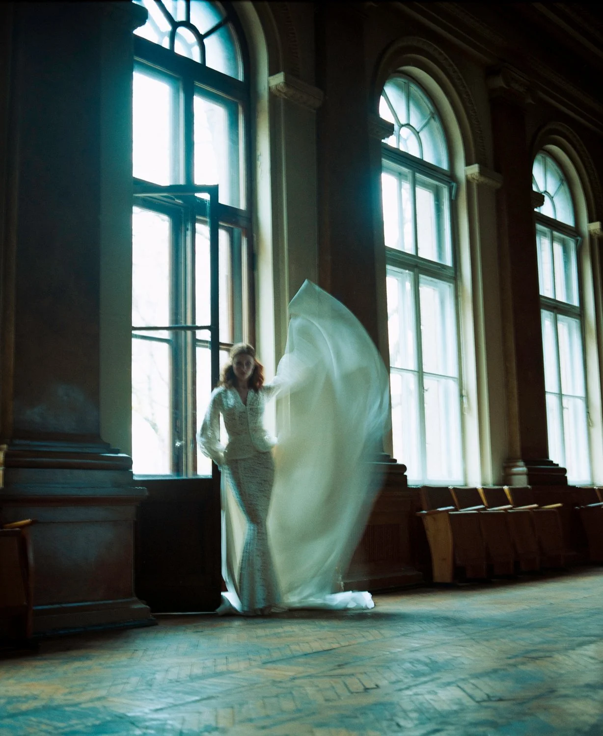 A woman in a white lace dress inside a spacious room with tall arched windows and wooden floors. She is standing near the window, with her hand on the wall, and a flowing piece of fabric or dress is caught in motion around her.