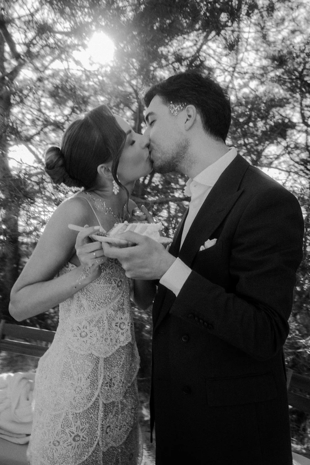A black and white photo of a bride and groom kissing outdoors, with trees in the background, as the groom holds a plate of cake.