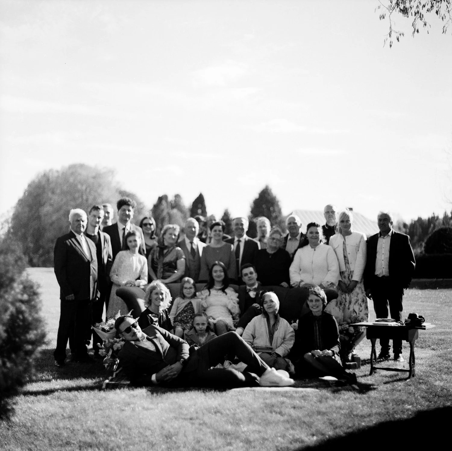 Black and white photo of a large group of people gathered outdoors on a grassy area for a group photo, some sitting and some standing, with trees and a building in the background.