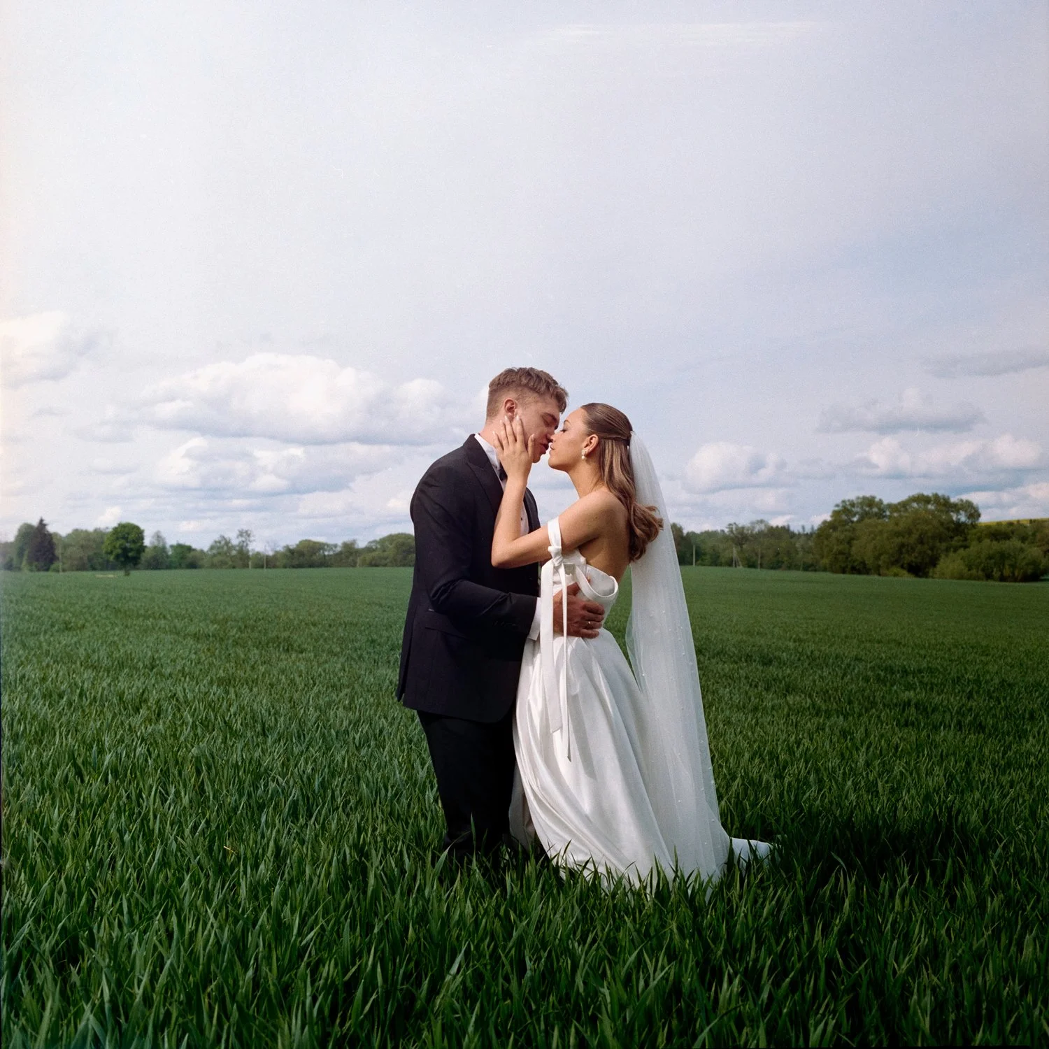 A bride and groom stand close on a green field with trees and cloudy sky in the background, sharing an intimate moment.
