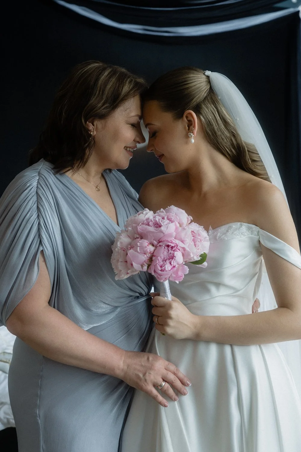 A bride and another woman, possibly her mother, share a moment with foreheads touching. The bride holds a pink bouquet and wears a white wedding dress and veil. The other woman wears a silver gown.