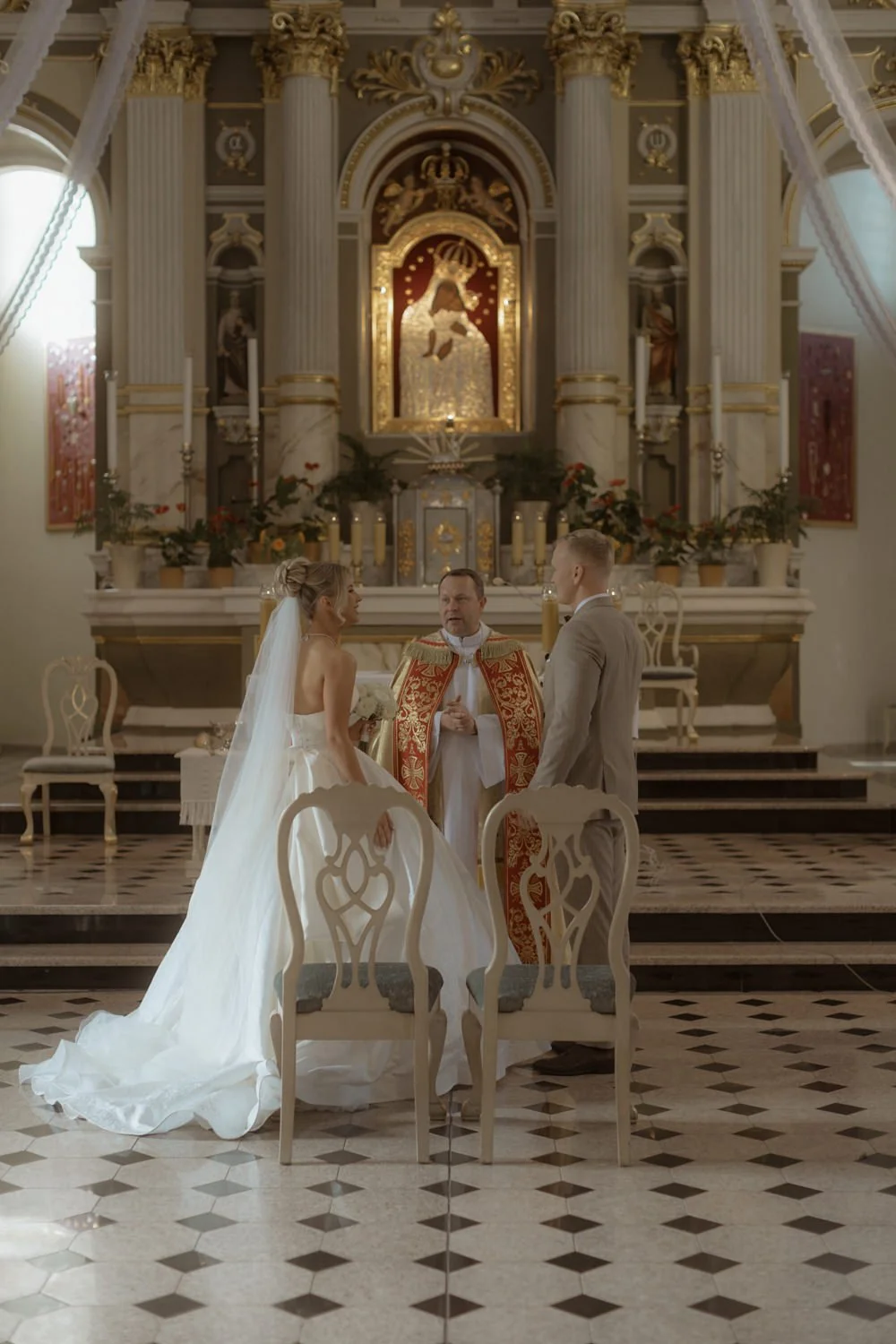 A bride and groom standing before a priest during a wedding ceremony in a church with ornate gold and white altar in the background.