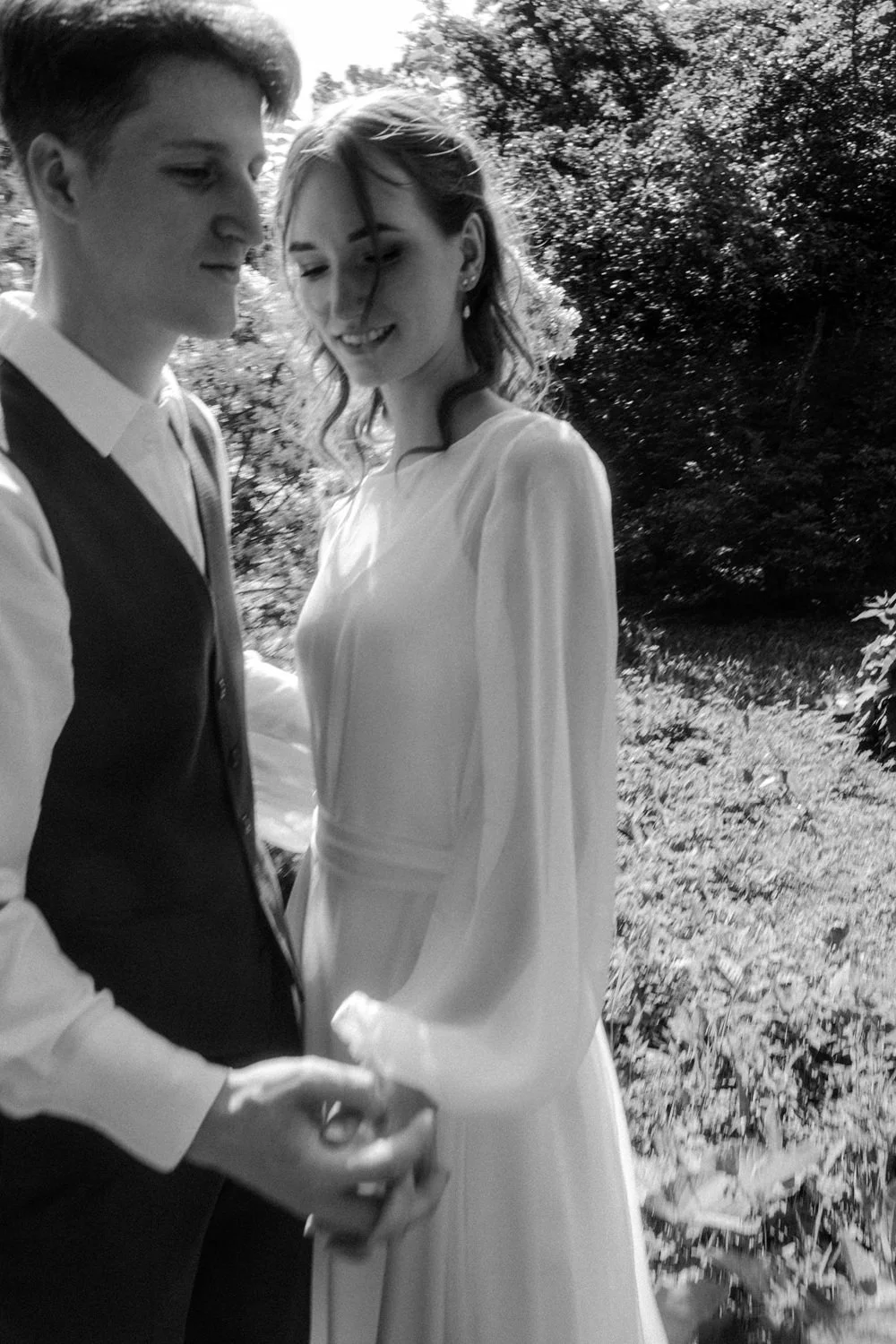 A black and white photo of a smiling couple holding hands outdoors with trees and plants in the background.
