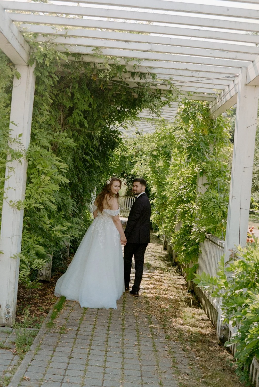 Bride and groom holding hands under a white pergola covered in green foliage.