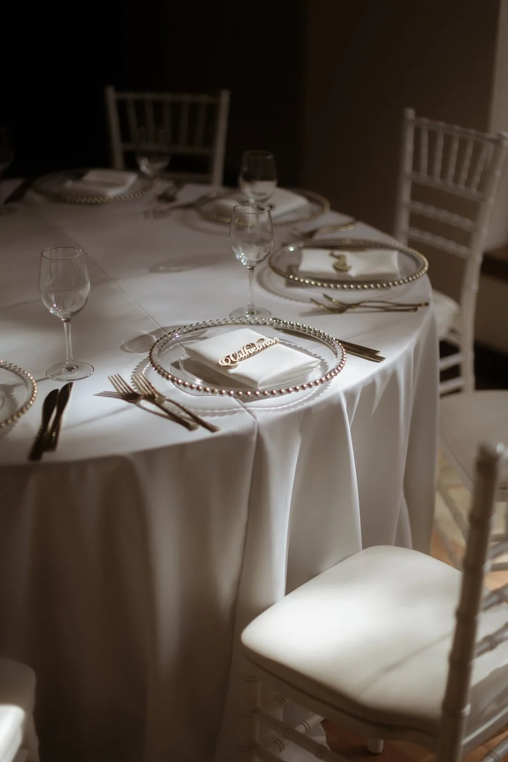 Elegant dining table set for a formal event with white tablecloth, beaded silver-edged plates, folded napkins, silverware, and empty wine glasses, illuminated by soft lighting.