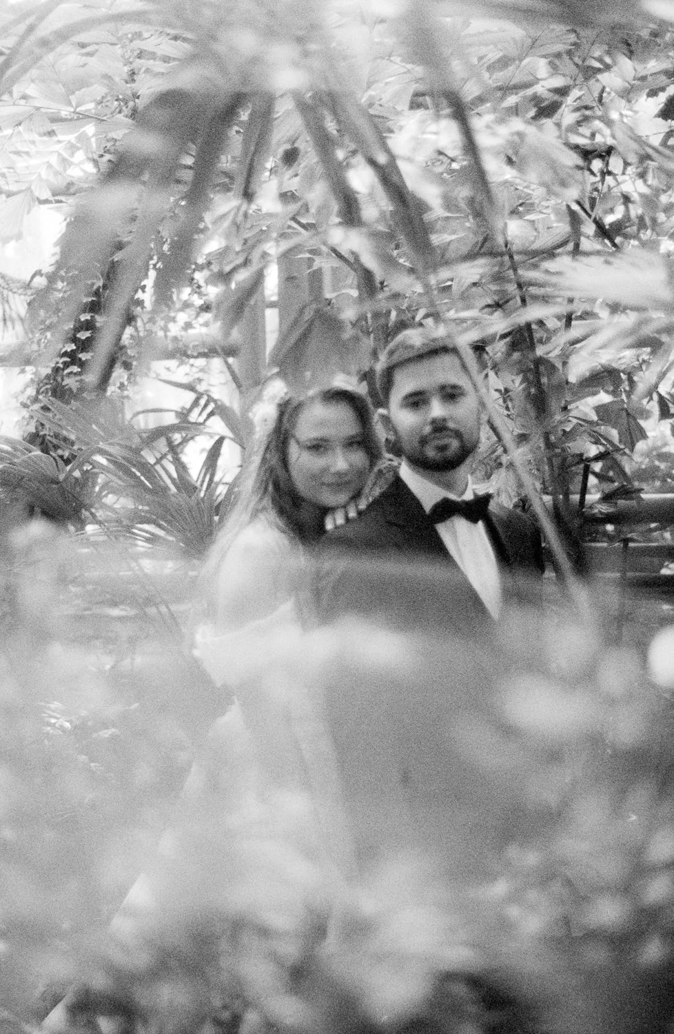 A black-and-white photo of a couple, a woman and a man in a tuxedo, among dense foliage and large leaves.