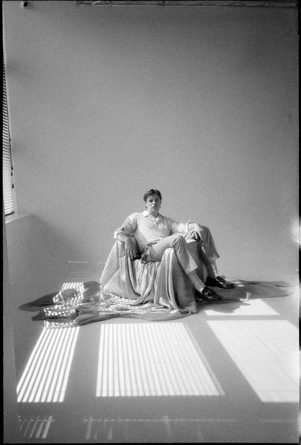A man sitting on a pile of draped fabric in a minimalist room with sunlight streaming through blinds, casting striped shadows on the floor.