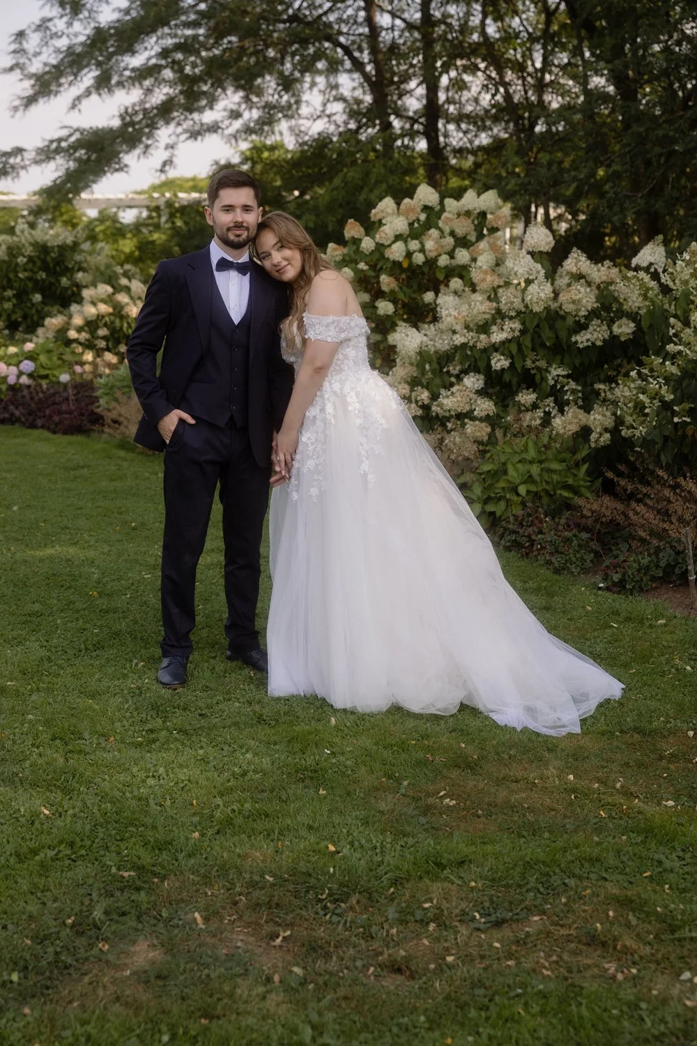 A couple in wedding attire standing on a grassy area with flowering bushes and trees in the background. The bride is leaning her head on the groom's shoulder, and they are holding hands.