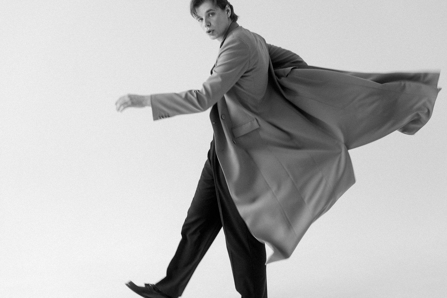 A young man in a long coat and dress pants is captured mid-motion, as if falling or spinning, against a plain background in a black and white photograph.