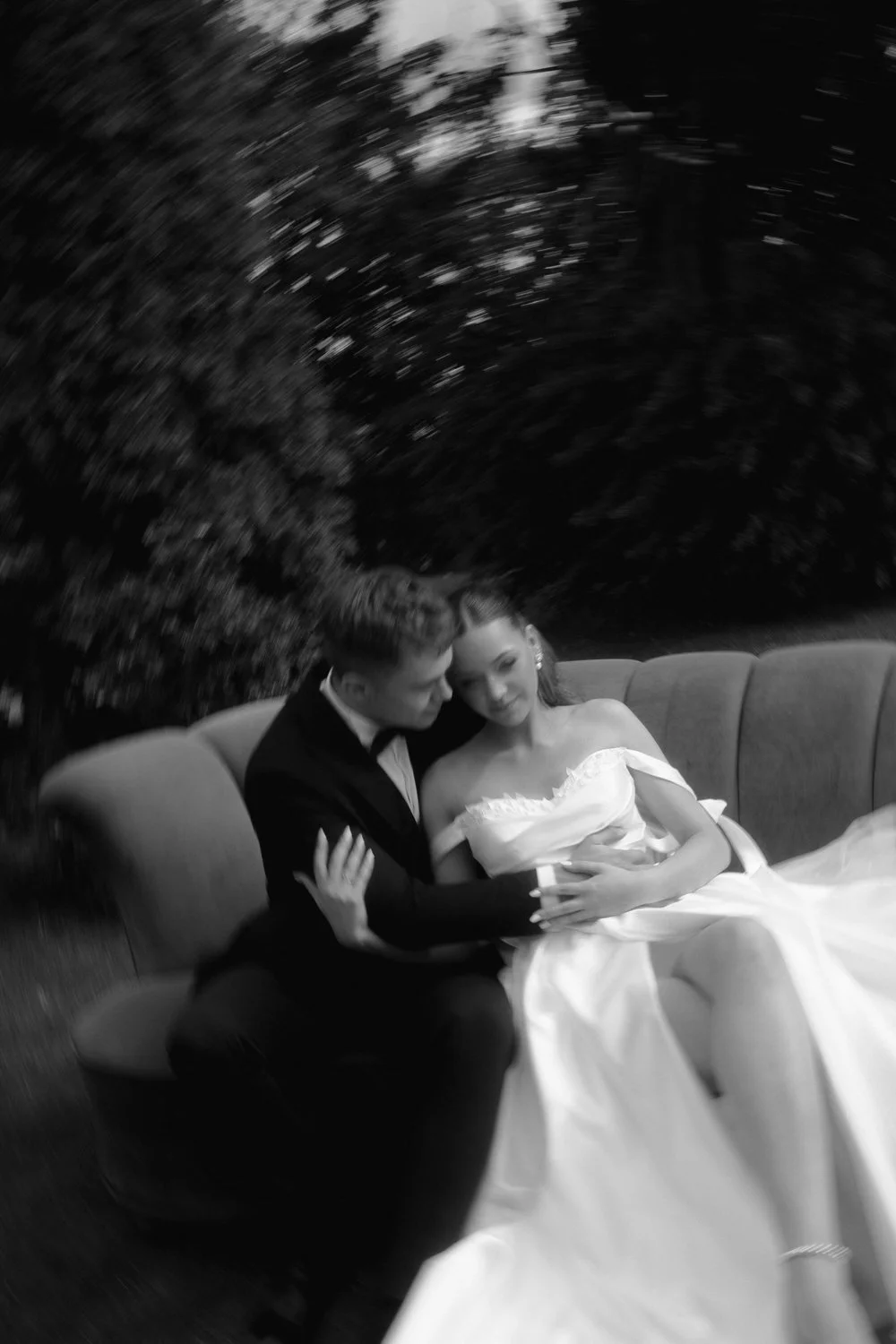 A black and white photo of a bride and groom sitting on a sofa, embracing each other and gazing into each other's eyes, outdoors with trees in the background.