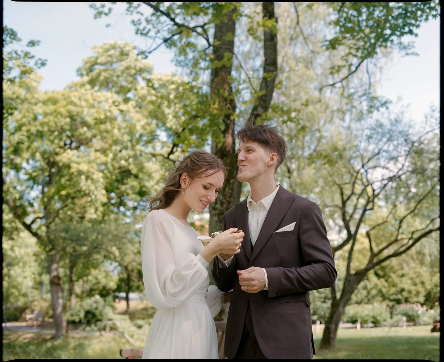 A young woman and man, dressed in white and black formal attire, standing outdoors under tall trees with green leaves. The woman is smiling and looking down at something she is holding, while the man is looking at her with a smile.