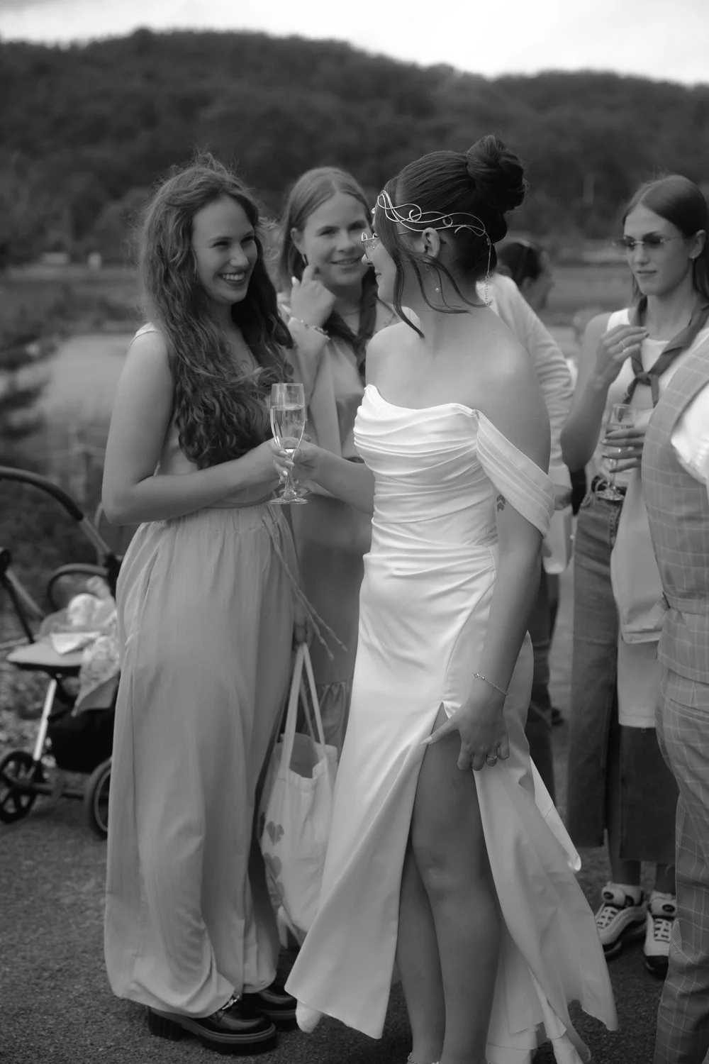 Group of women at a celebration, one woman in a white dress talking to others, holding a glass of champagne, outdoors with hills in the background.