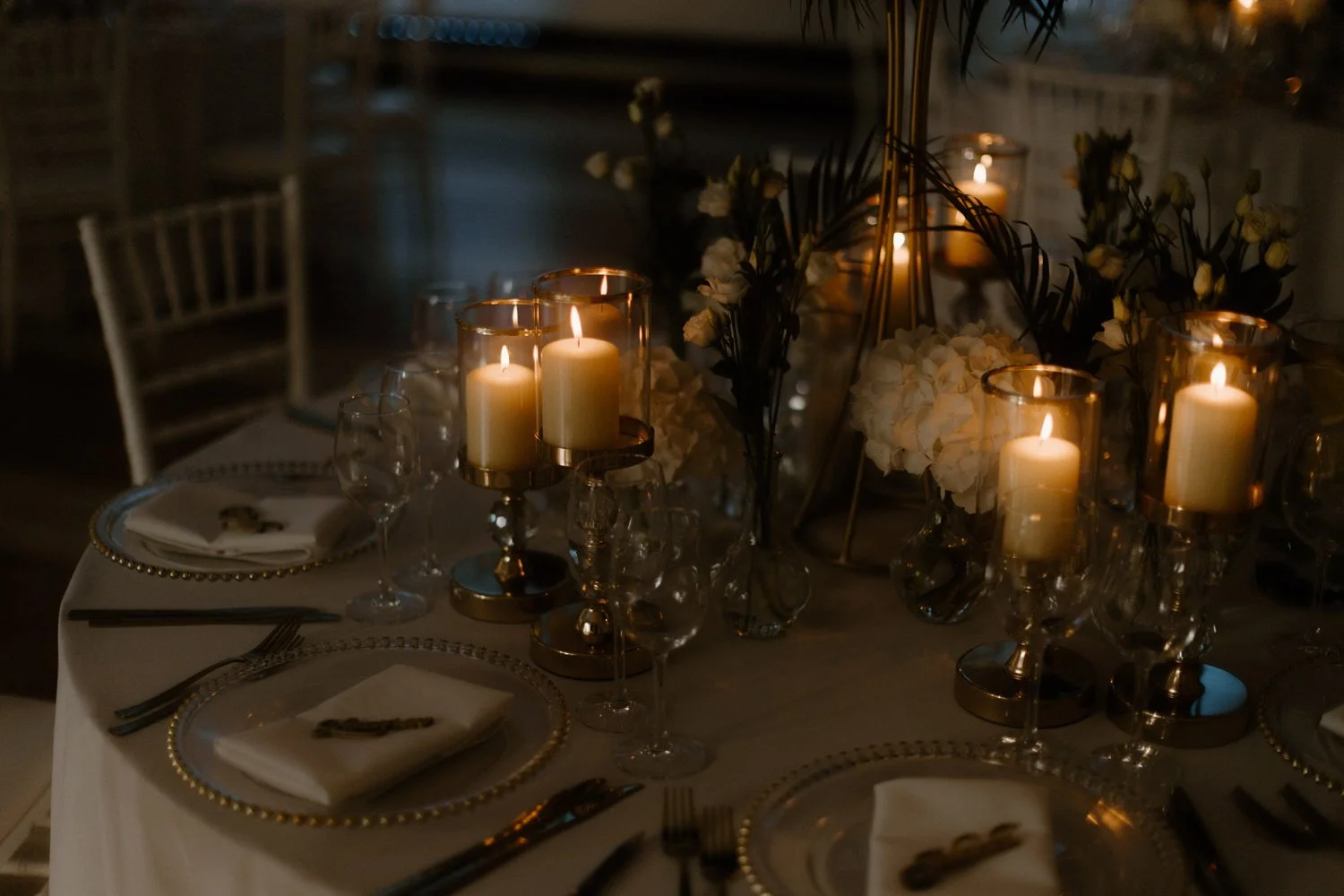 A table set for a formal event with candles, glassware, and floral centerpieces in a dimly lit setting.