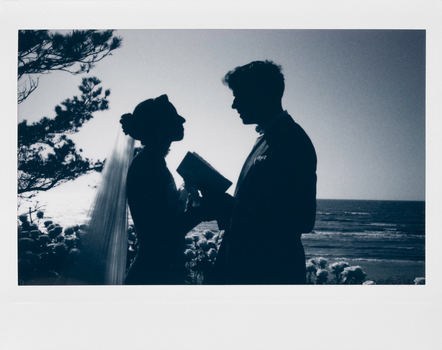 Silhouettes of a bride and groom standing by the ocean at sunset, with the bride holding a bouquet and the groom reading from a book or vows.