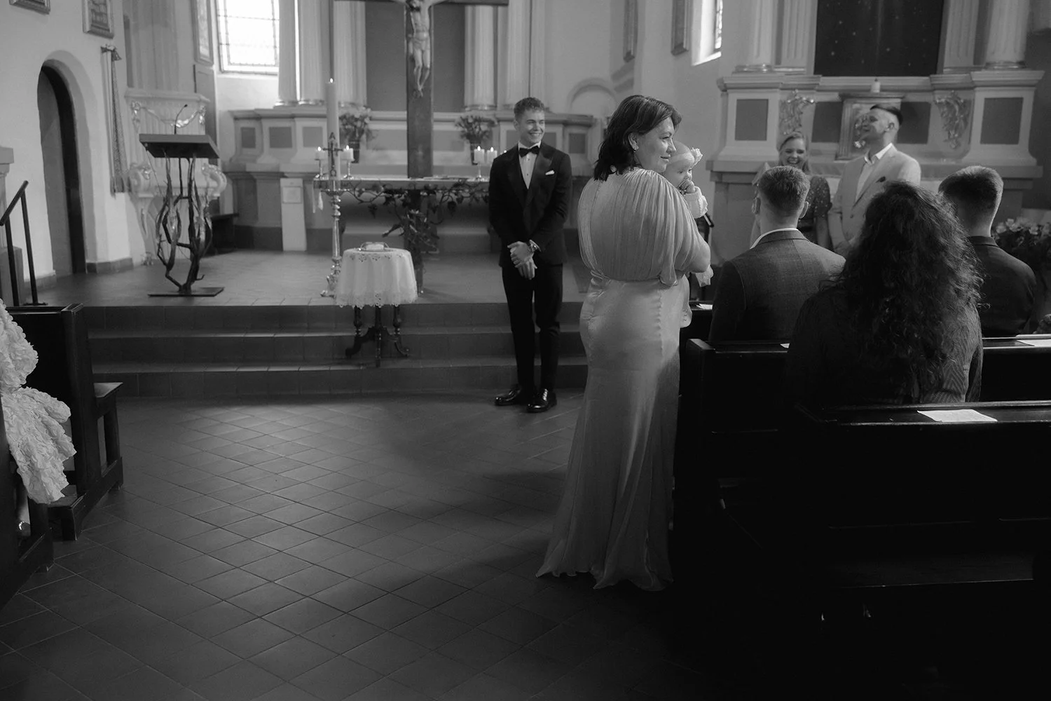 Black and white photo of a wedding ceremony inside a church, with the bride holding a baby, standing near the altar, and the groom in a suit standing nearby. Several guests are seated in pews, facing the front, during the service.
