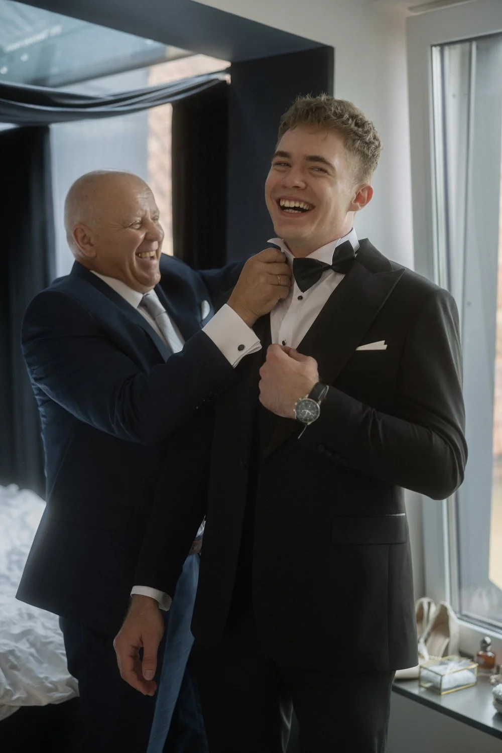 An older man in a navy suit helping a young man in a tuxedo adjust his bowtie, both smiling happily in a well-lit room.