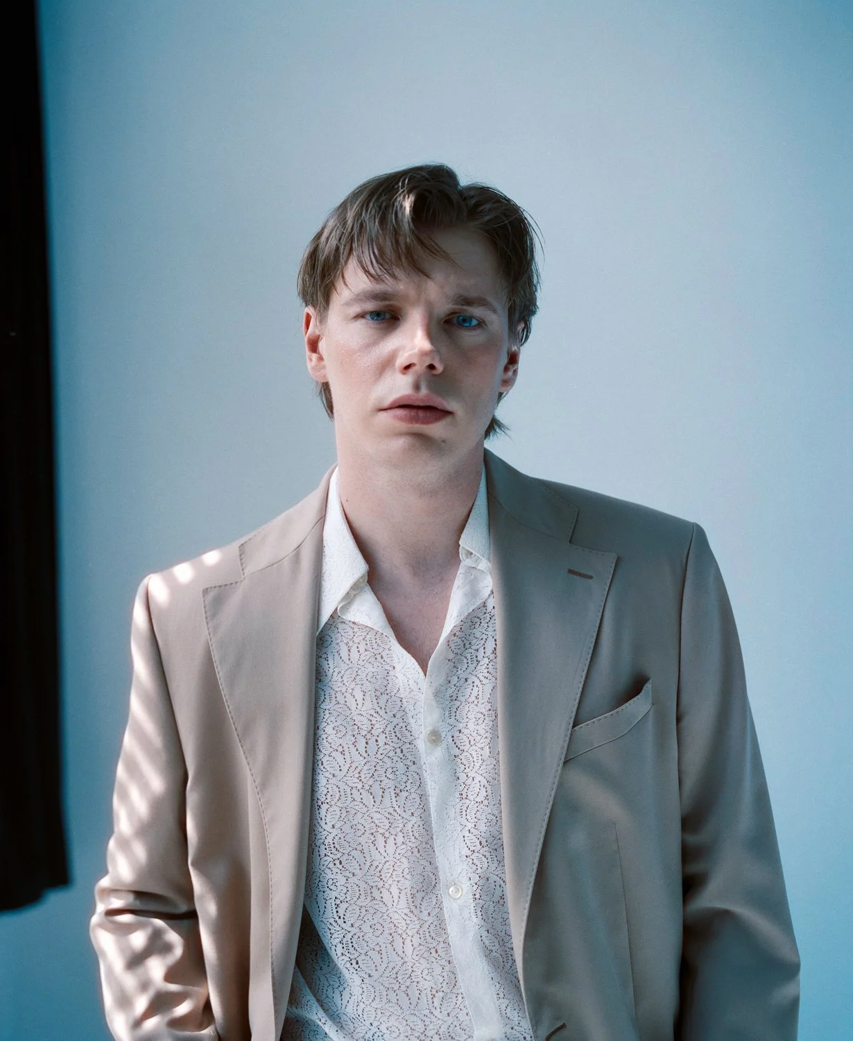 A young man dressed in a beige suit and white shirt standing against a blue wall.