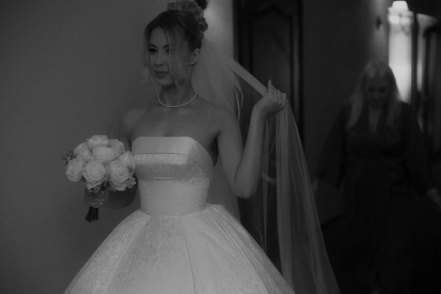 A bride in a strapless wedding gown holding a bouquet of roses, with her veil being adjusted by someone in the background.
