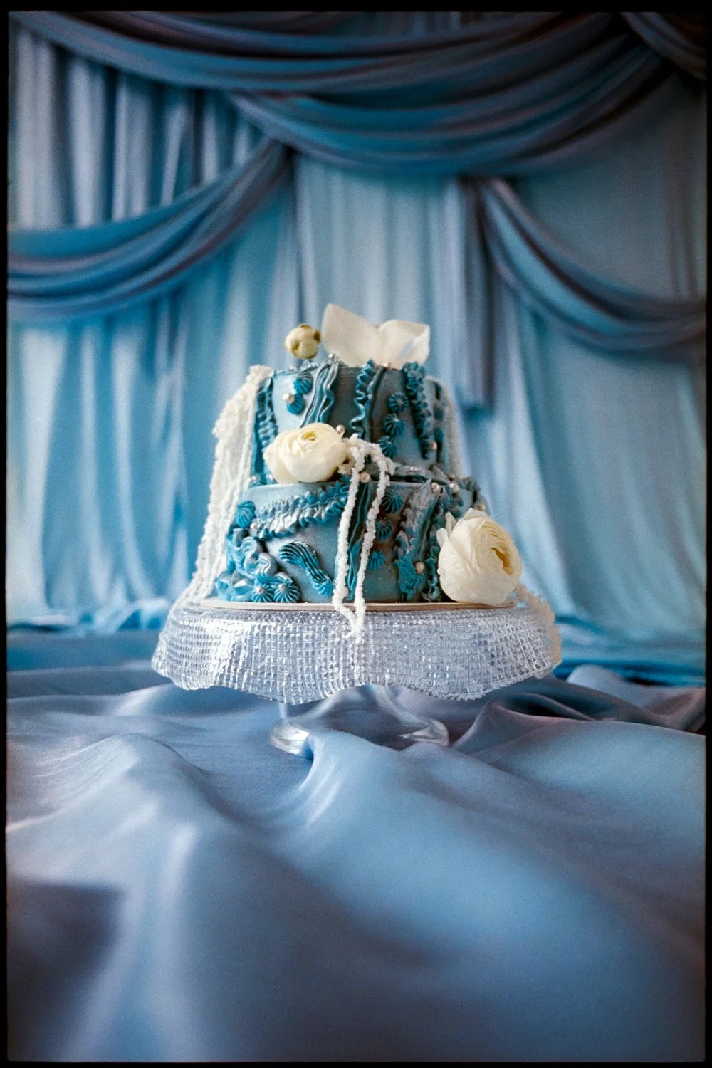 A wedding cake decorated with blue and white icing, white flowers, and detailed piping, displayed on a stand with a white textured cloth, set against a backdrop of blue drapes.