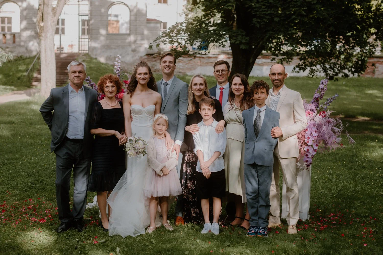 Group of fifteen people, including children, adults, and seniors, posing outdoors on a lawn with trees and a building in the background, dressed in formal attire for a celebration or wedding.