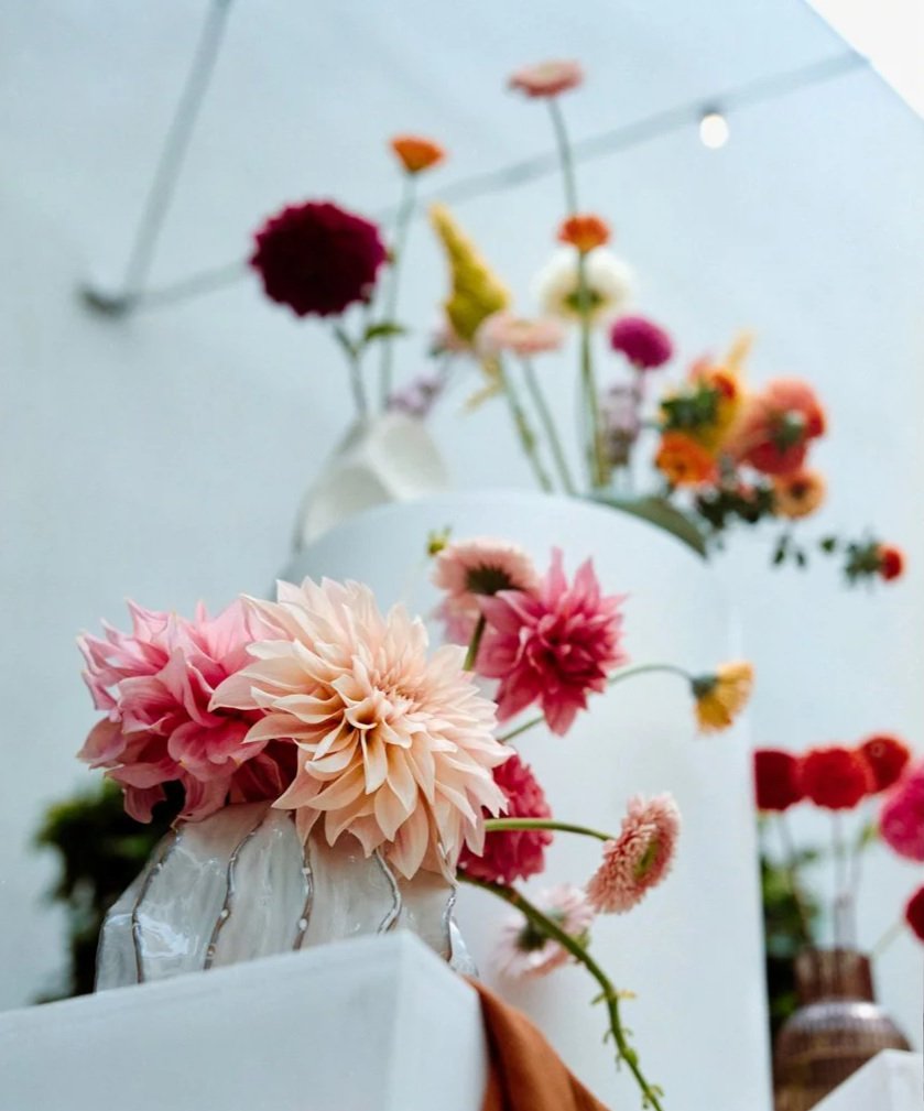 Vase with pink and peach dahlias and other colorful flowers, set on a white surface with a light blue background and ceiling support visible.