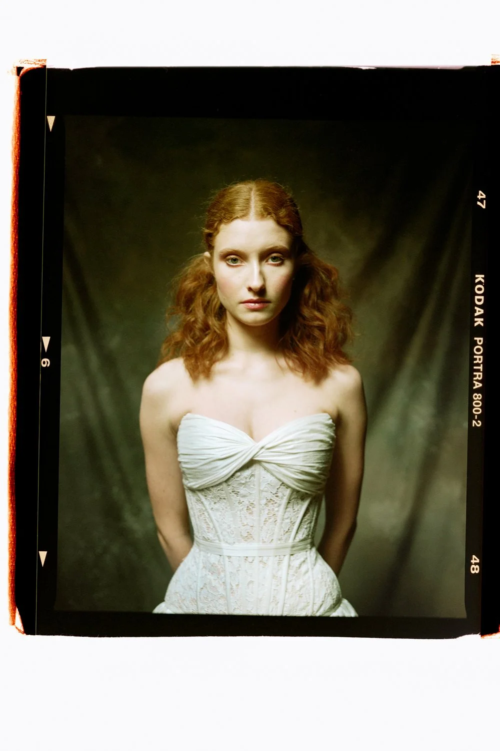 A young woman with red, curly hair styled in loose waves, wearing a strapless white dress with lace details, standing against a dark, textured background.