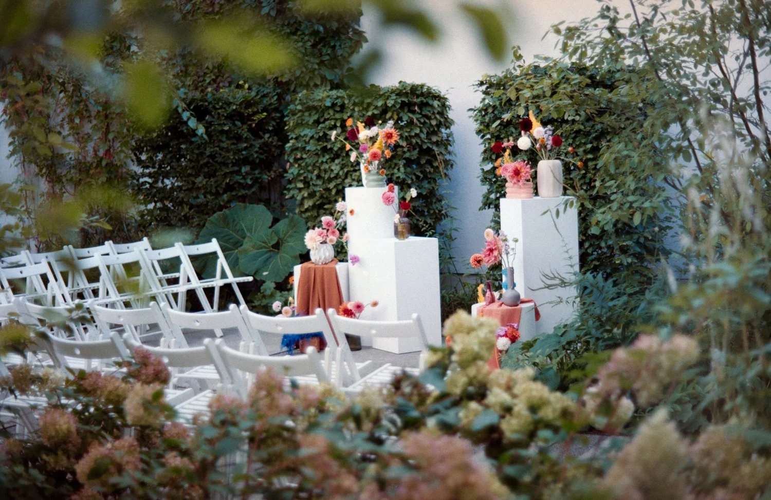 Decorative floral display with white chairs, floral arrangements, and pedestals in an outdoor garden setting, likely prepared for an event.
