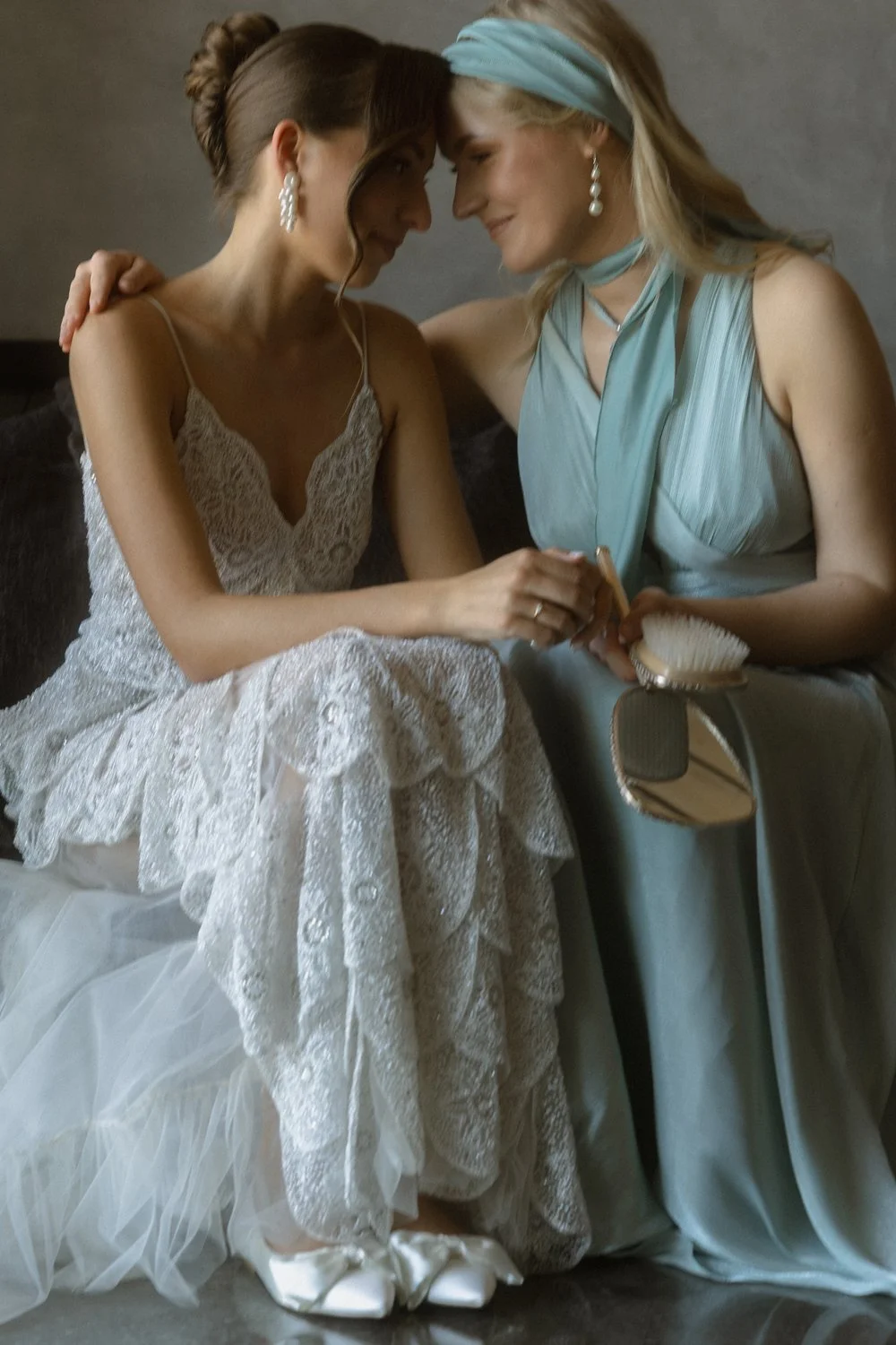 Two women sharing an intimate moment, with foreheads touching and eyes closed. One is dressed in a wedding gown with lace detailing, and the other is dressed in an elegant light blue dress with matching headscarf and earrings.