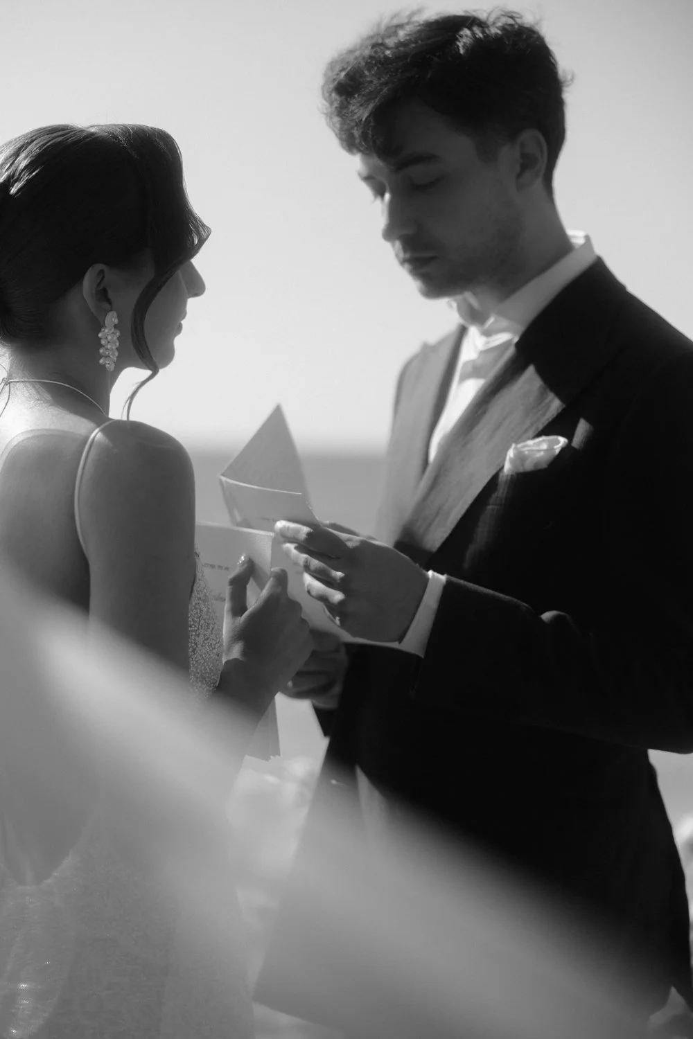 A black and white photo of a wedding ceremony, with a bride and groom exchanging vows and holding hands, the bride wearing earrings and a dress, and the groom in a suit.